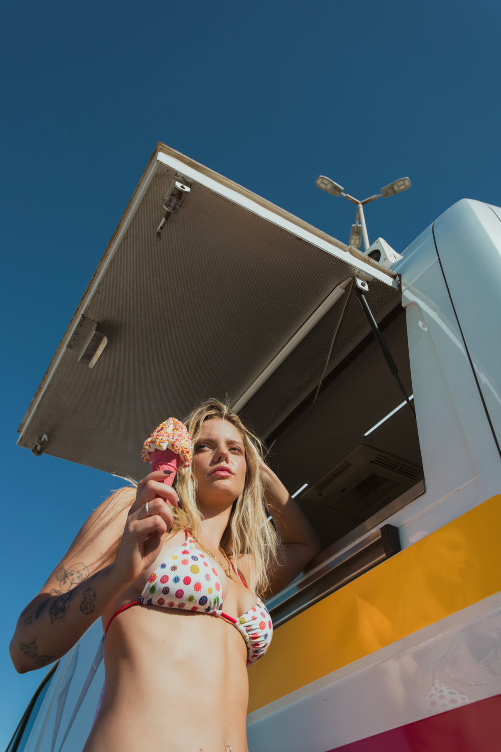 A woman in a bikini stands confidently on the shore, with a clear blue sky above.